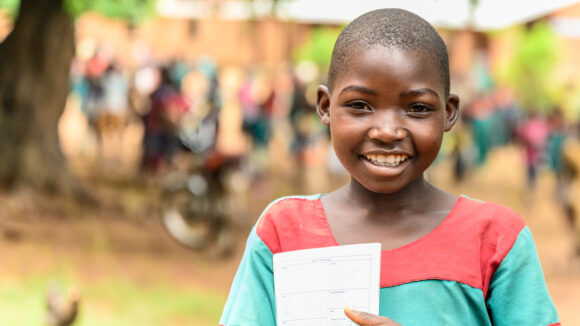 Mary, a ten-year-old girl, stands outside while holding her school exercise book and smiling.