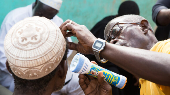 An eye health worker uses a torch while checking a man's eyes for trachoma.