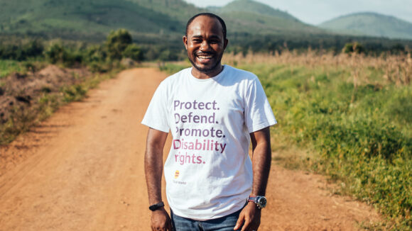 Sightsavers researcher Martins smiles as he stands in the middle of a road. He's wearing a Sightsavers t-shirt that reads 'Protect. Defend. Promote. Disability rights.'