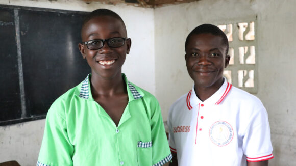 Twelve-year-old Arthur smiles as he stands next to his teacher, Mr Thompson. They stand in front of a chalk board in a classroom.