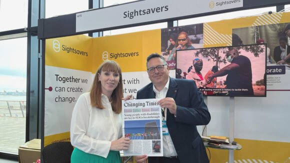 Deputy prime minister Angela Rayner and Sightsavers' Ross Gilligan stand together holding a copy of Sightsavers' Changing Times newspaper. They are standing in front of Sightsavers' stand at the Labour party conference.
