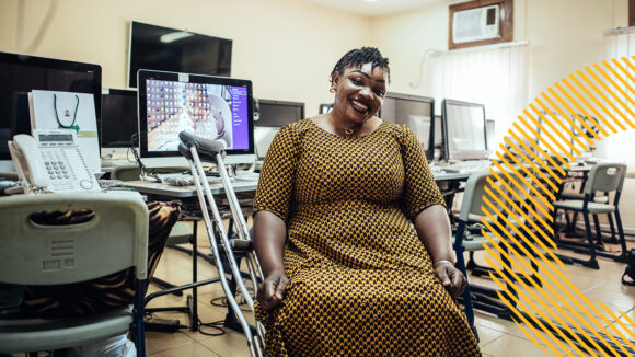 Onyinyechi, a polio survivor, works as an information analyst. She is seated in front of a computer, smiling in her yellow and black dress, with crutches leaning against her desk.