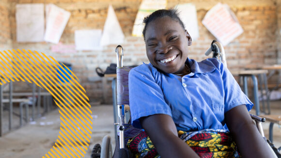 A girl smiles in a classroom in Zambia. She's sitting in a wheelchair and wearing a blue school shirt.