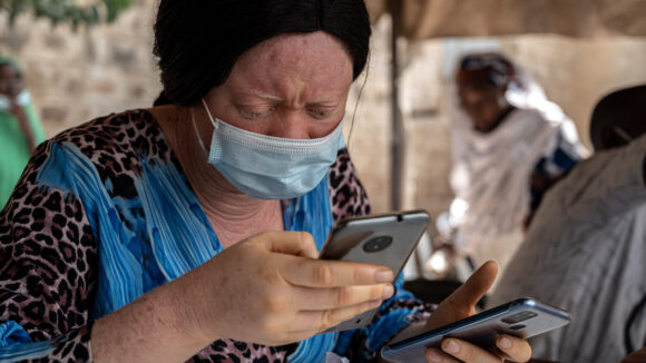 A woman wearing a surgical face mask holds two mobile phones while recording data about cataract operations in Nigeria.