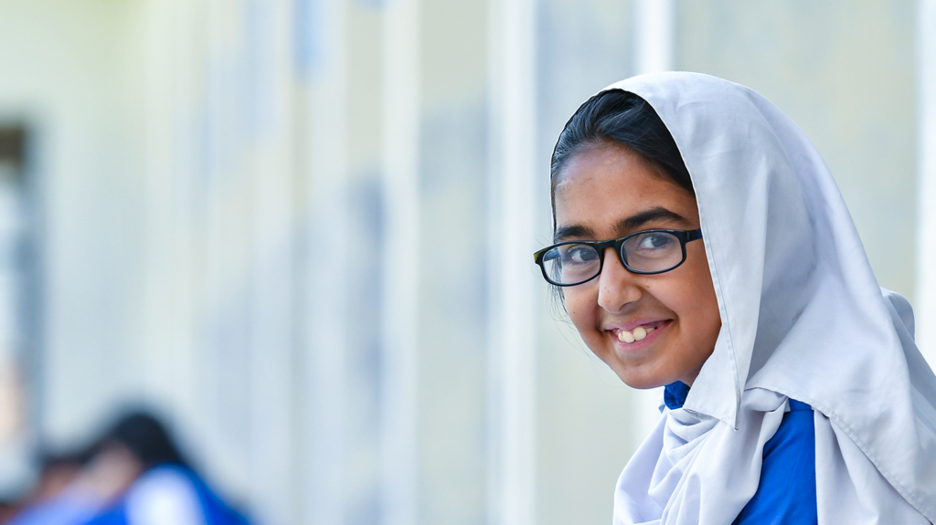 A girl, who wears glasses, smiles as she sits in a school corridor.
