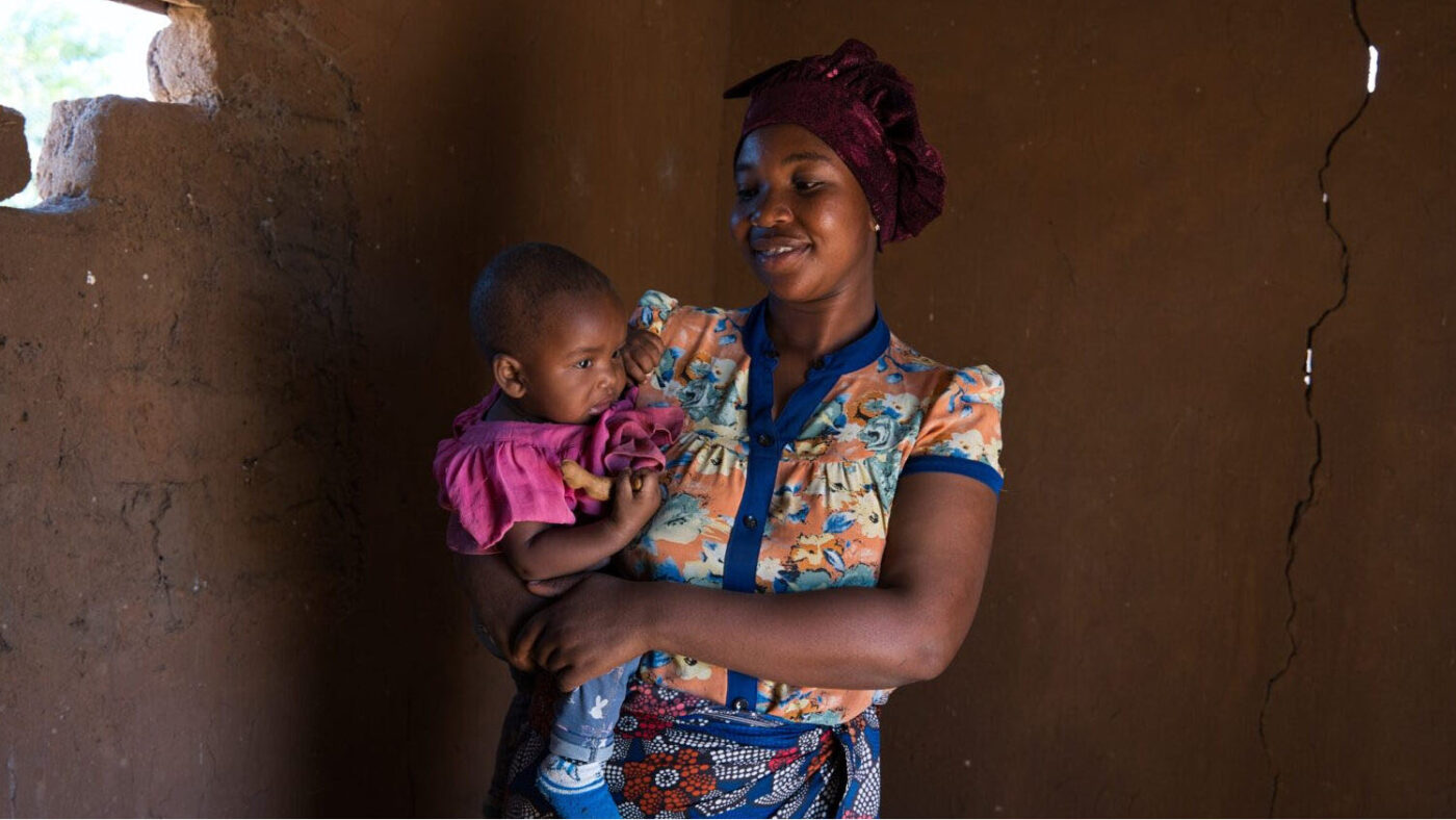 Rudia holds her daughter, Ebenezeri, inside her home. She's smiling and looking at Ebenezeri.