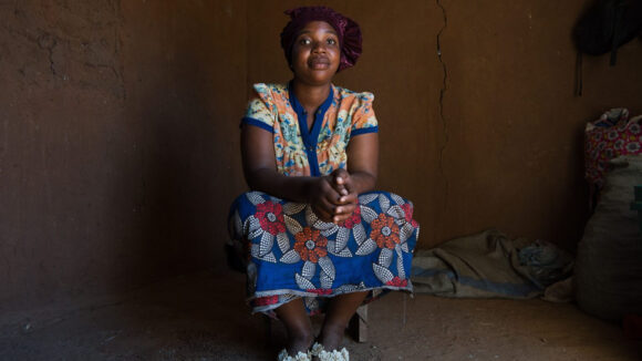 Rudia sits inside in her home and looks at the camera.