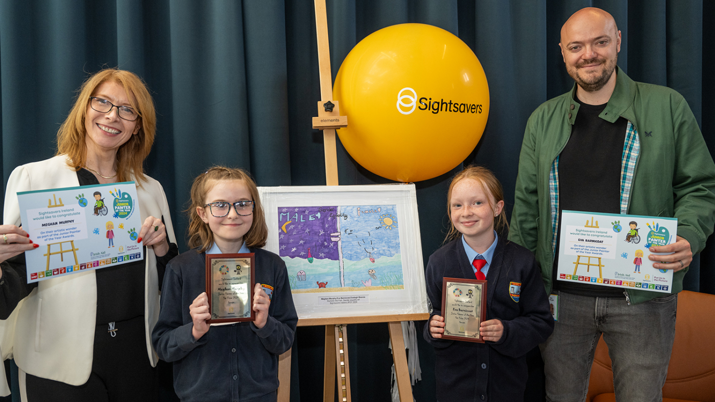 Two girls holding trophies and two members of staff stand beside a painting on an easel.