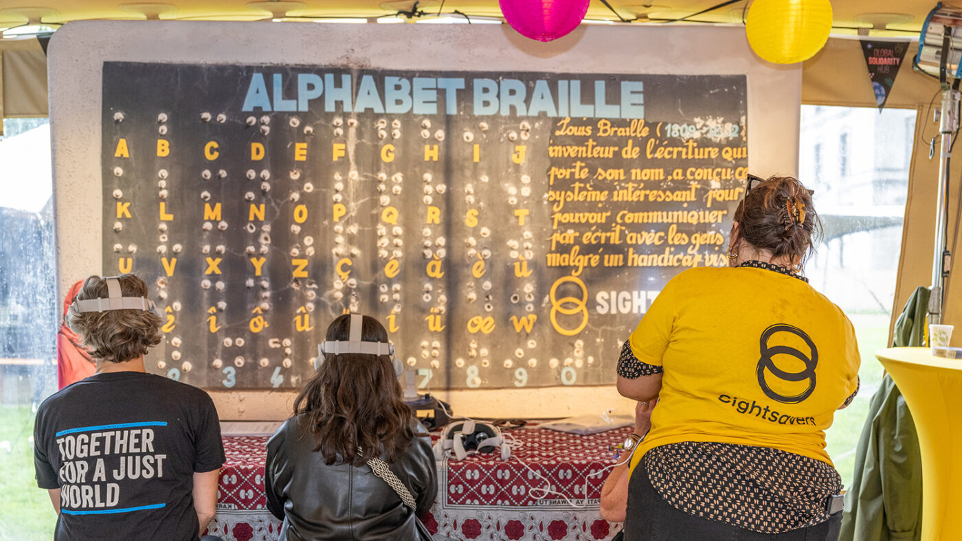 Two women wear virtual reality headsets while sitting in front of a poster that shows the alphabet in Braille.