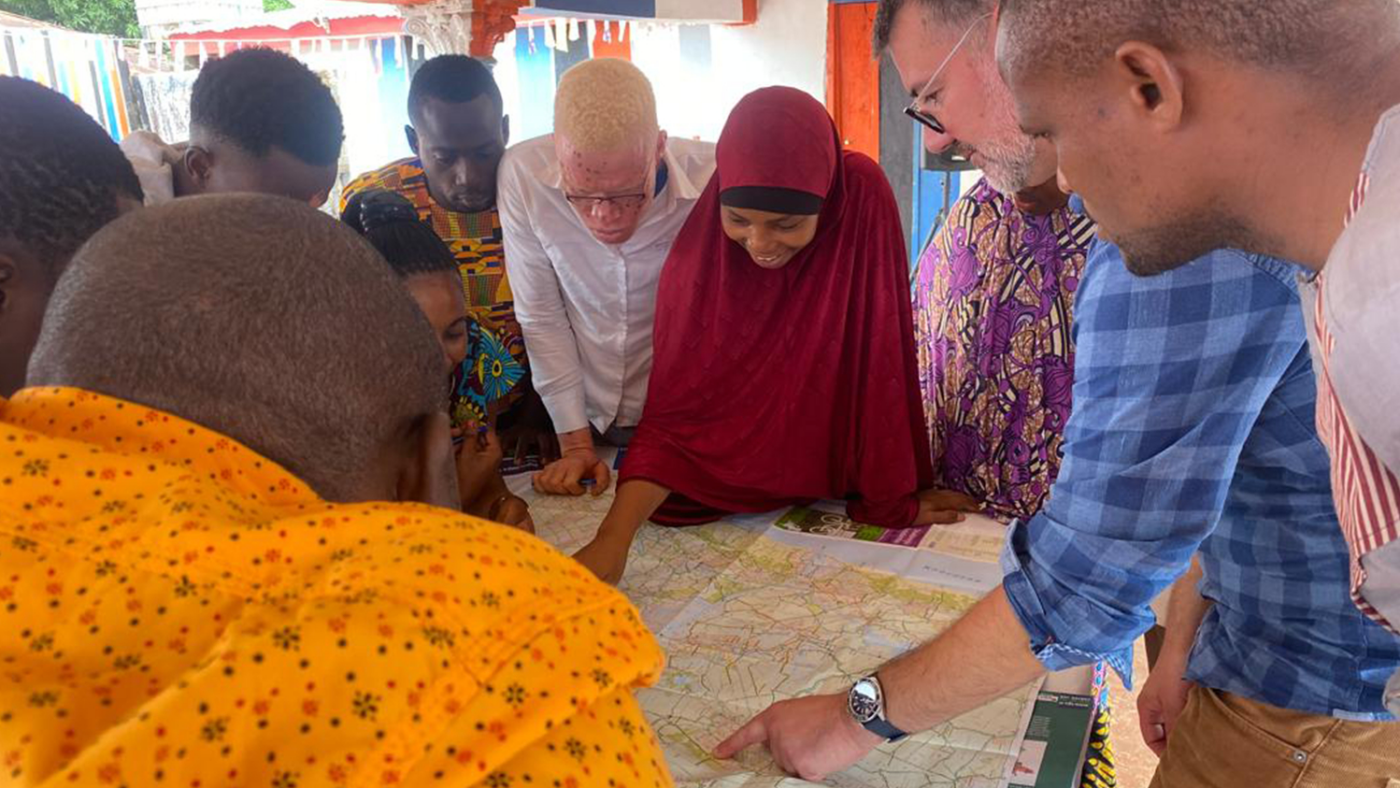A group of young researchers gathers around a table, watching as an adult points to a map.