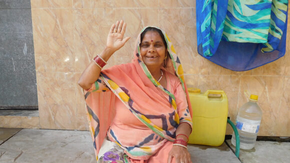 A middle-aged woman wearing a sari smiles and waves.