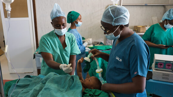 In an operating theatre, a surgeon and a nurse prepare baby Ebenezeri for her cataract operation. The medical staff are wearing green scrubs and surgical masks.
