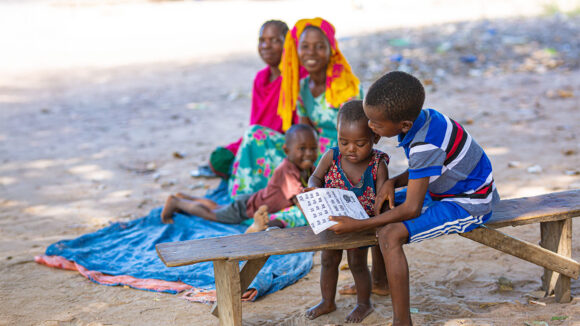 Mohamed sitting on a bench, reading to his younger cousin as his family watch on.