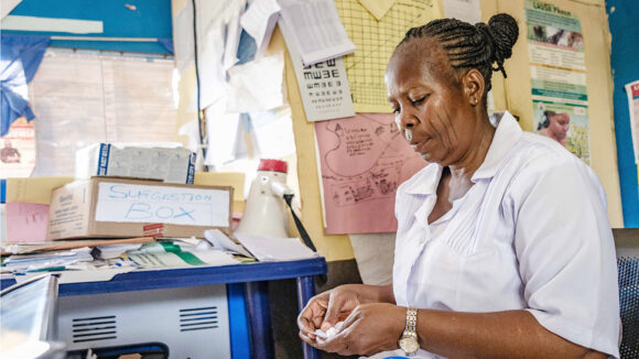 Health worker Adesugba checks some medication in the clinic. She's surrounded by charts on the walls and paperwork on the tables.