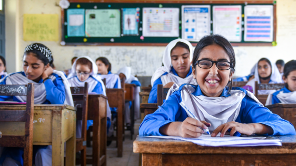 Minahil, who wears spectacles, smiles broadly as she sits at her classroom desk.