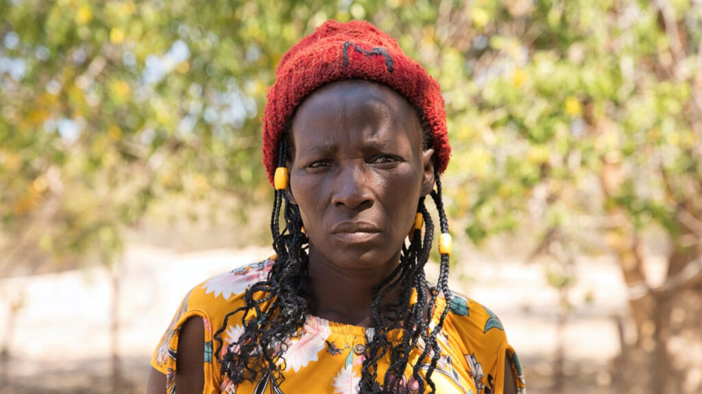 Maria wears a red knitted hat and a yellow floral shirt, stood in front of trees.