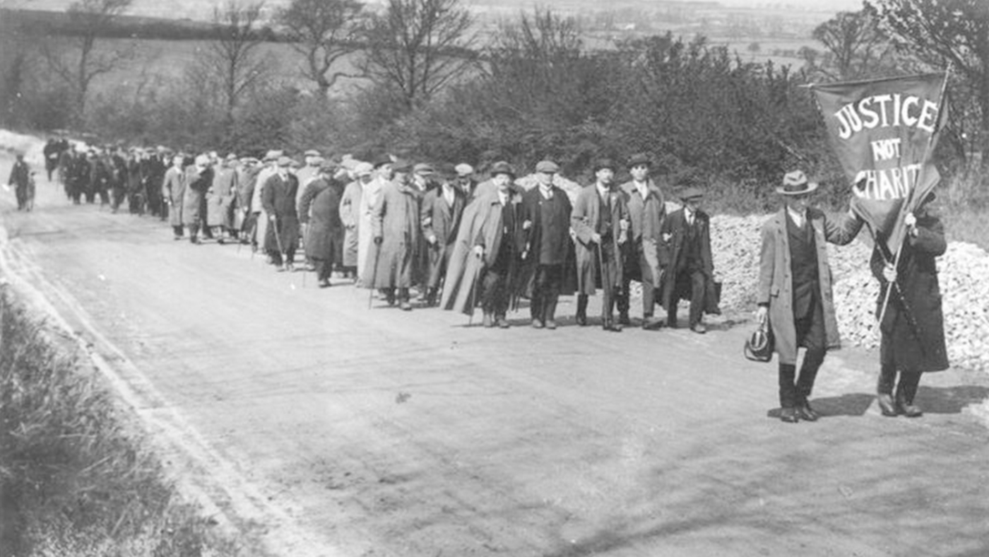 A large group of men march down a country road with a banner reading ‘justice not charity’ at the head.