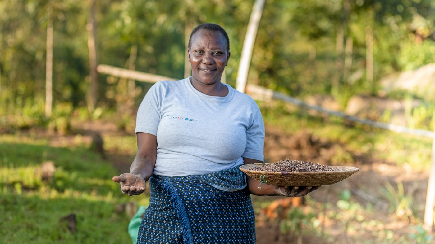 A woman stands in a field holding a basket of sorghum in one hand and in her other, holds the crop towards the camera.