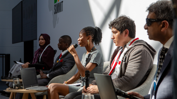 Four panellists listen to a woman speaking into a microphone. One man wears sunglasses for a visual impairment.