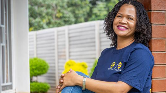 Samantha Nyathi, who is wearing a Sightsavers-branded T-shirt, smiles while leaning against a wall in a garden.