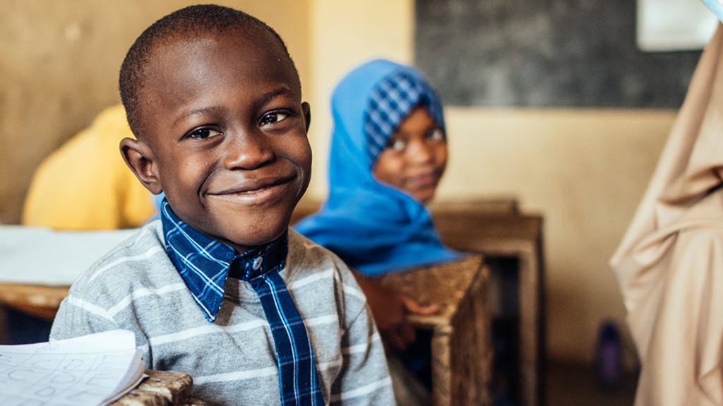 A portrait of Muhammad smiling, sat at his desk in school.