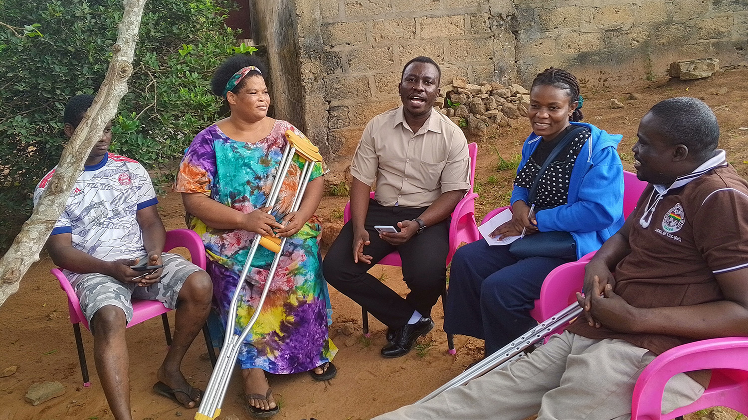 A group of five people in conversation sit in a semicircle under a tree during a qualitative interview.