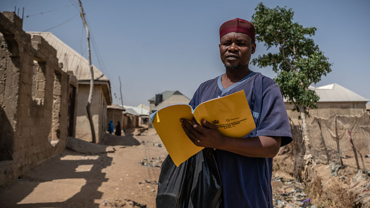 Standing outside some houses, Aliyu writes in a yellow Sightsavers notebook.