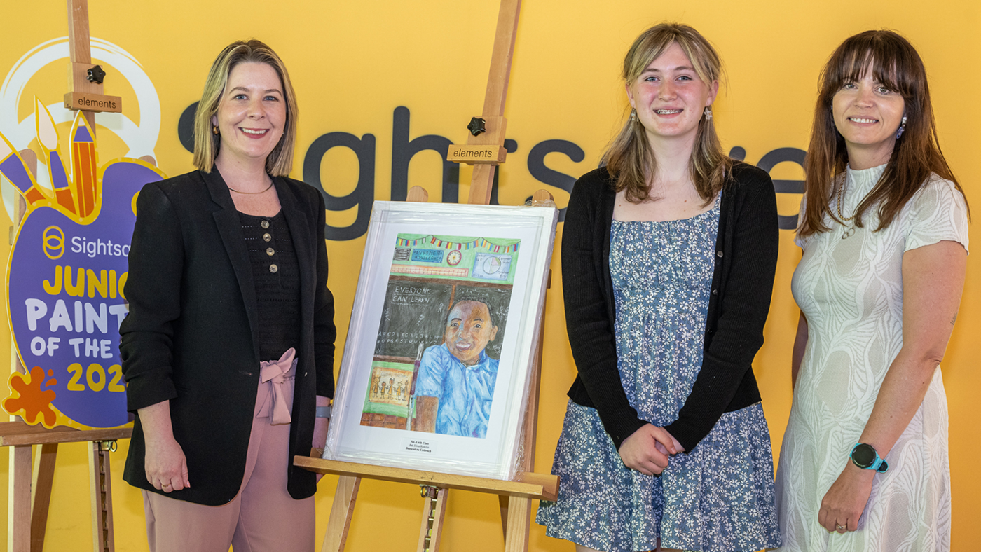 Two women and a young adult stand beside a canvas on an easel.
