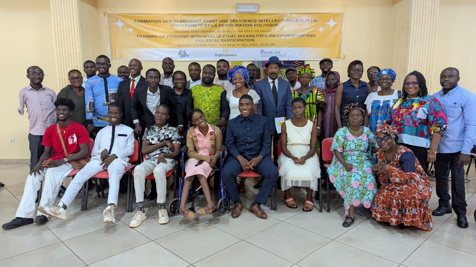 A large group of men and women pose for a photo. Behind them is a banner that reads 'Training of persons with intellectual disabilities on citizenship and political participation'.