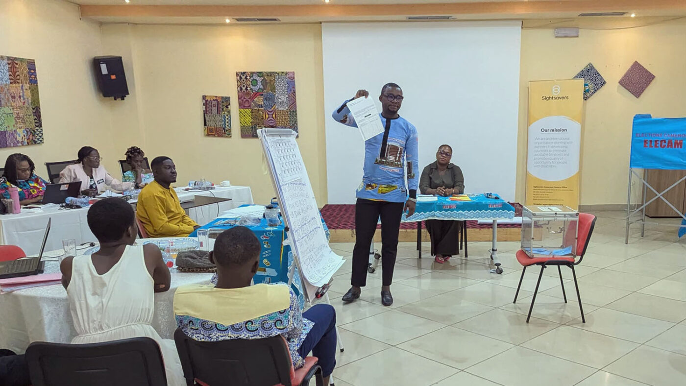 A workshop leader stands at the front of a room holding up a sheet of paper.