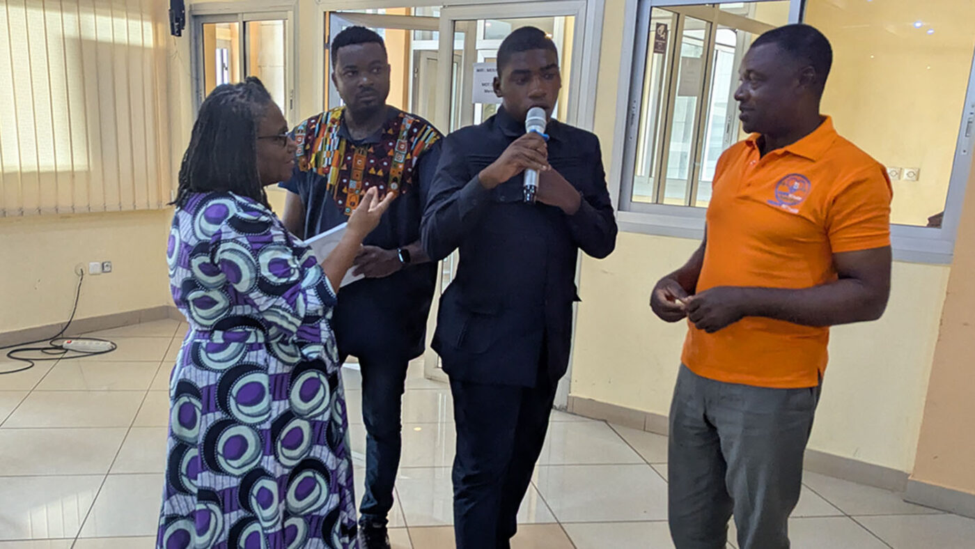 A young man with intellectual disabilities speaks into a microphone. Workshop facilitators stand on either side of him.