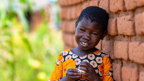 Eight-year-old Mwamini smiles after her successful operation to treat advanced trachoma. She is leaning against a brick wall in her village.