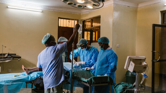 A surgeon performs an operation to treat advanced trachoma on Mwamini. Mwamini is lying on an operating table surrounded by four health workers wearing medical scrubs.