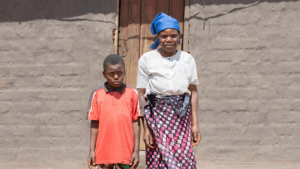 Chisomo and his mother Margaret stand in front of the wooden door of their house.