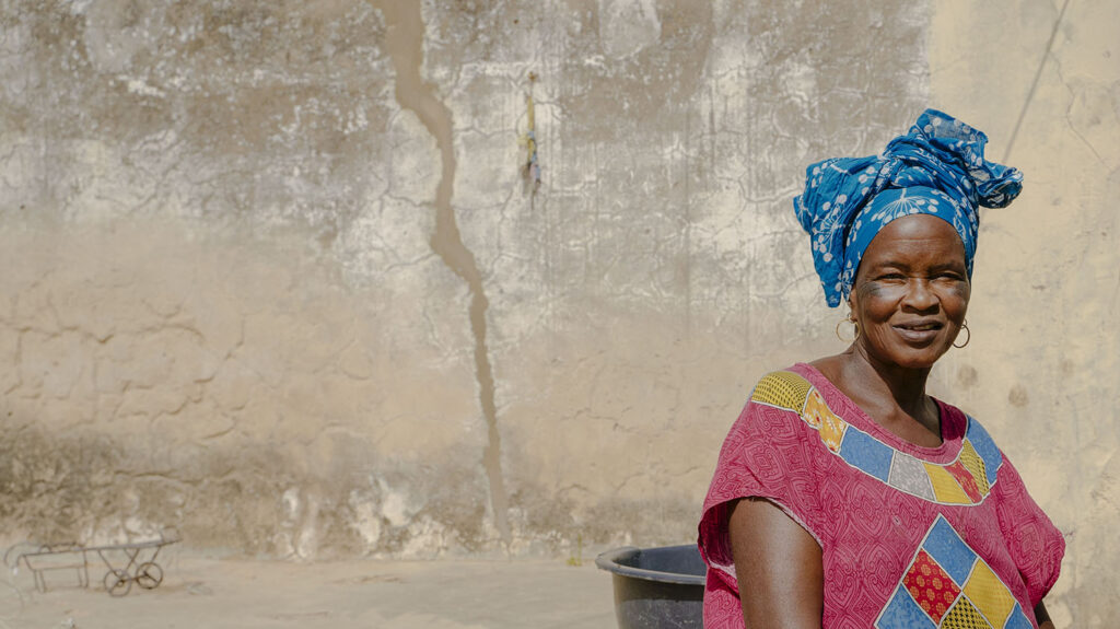 58-year-old Ndack, who had advanced trachoma, smiles after recovering from surgery for the eye condition. She's sitting outside a house, wearing a blue headwrap.