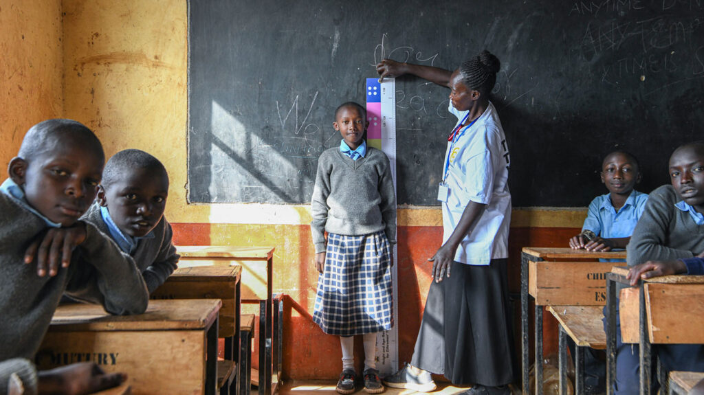 At school, a female community volunteer measures a girl to see how much medication she needs to protect her from trachoma. Other students sit at their desks in the classroom.