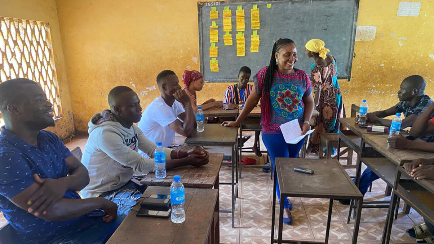 A woman stands in the middle of a classroom. People are sitting at desks around her.
