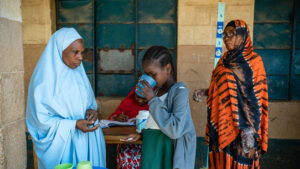 A young girl drinks from a cup after taking medication. Two women wearing burkas watch her.