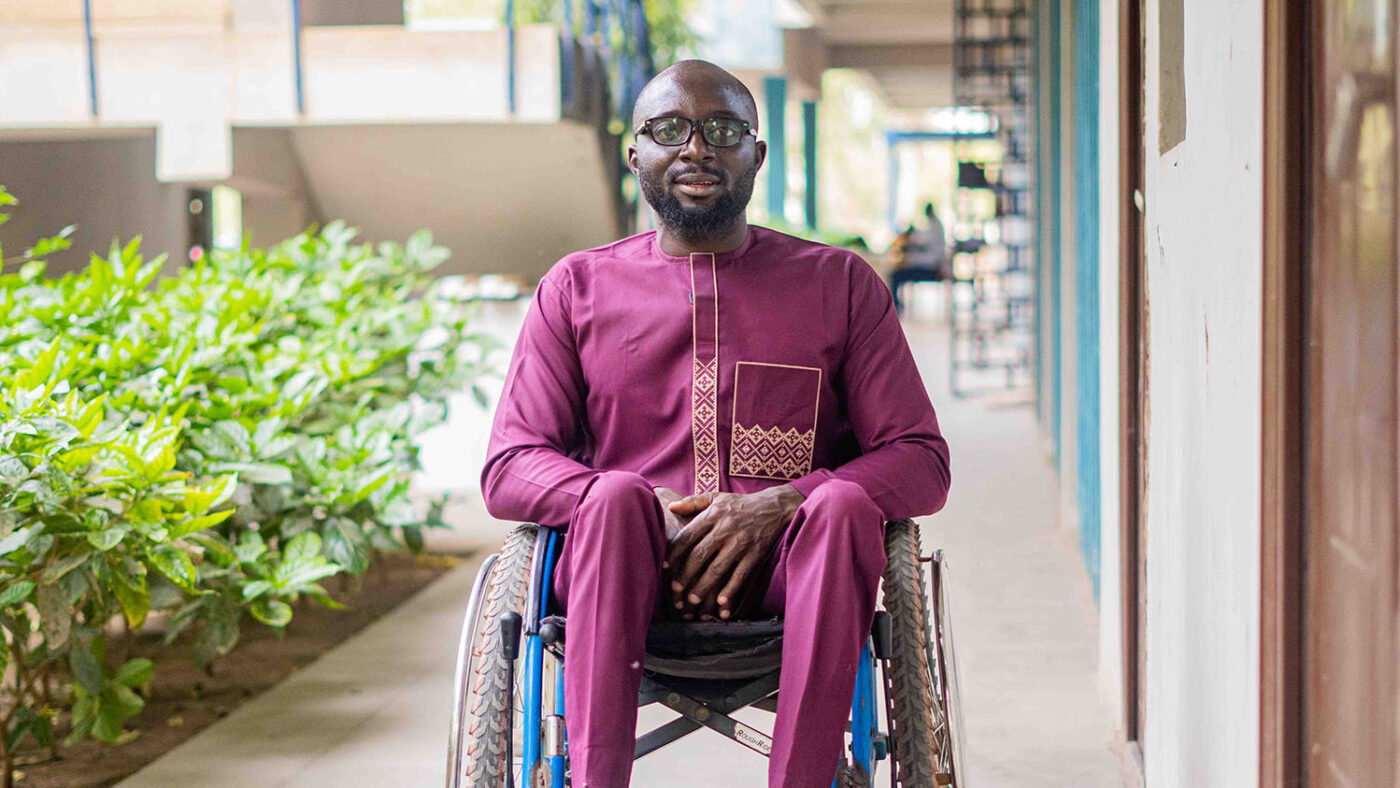 A man sitting in a wheelchair wearing a purple tunic and a pair of glasses.