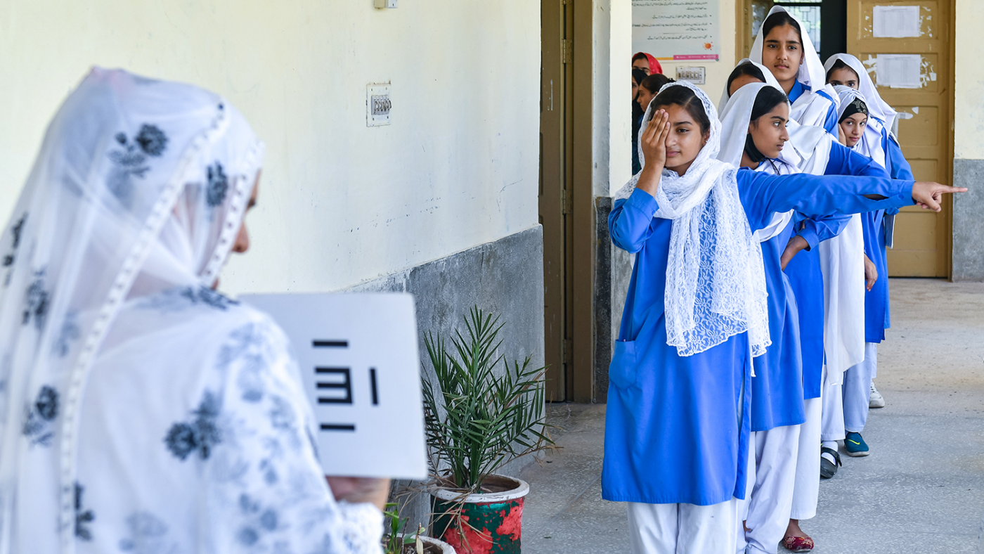 Girls in school uniform line up to get their eyes tested by a woman who holds an eye chart.