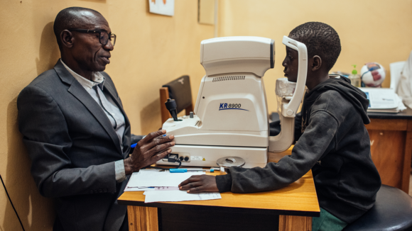 A young boy sits in a chair while a man in a suit examines his eyes using optometry equipment.