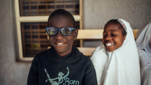 A boy wearing glasses smiling. Behind him, a girl wearing a hijab is beaming.