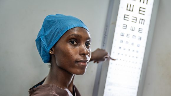 A female eye health worker points to a letter on an eye test chart. She is wearing a blue surgical scrub cap.