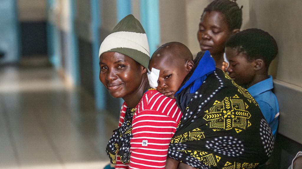 Felister sits in a hospital corridor with Chancy on her back in a baby sling wrap. Chancy has a bandage over his right eye.