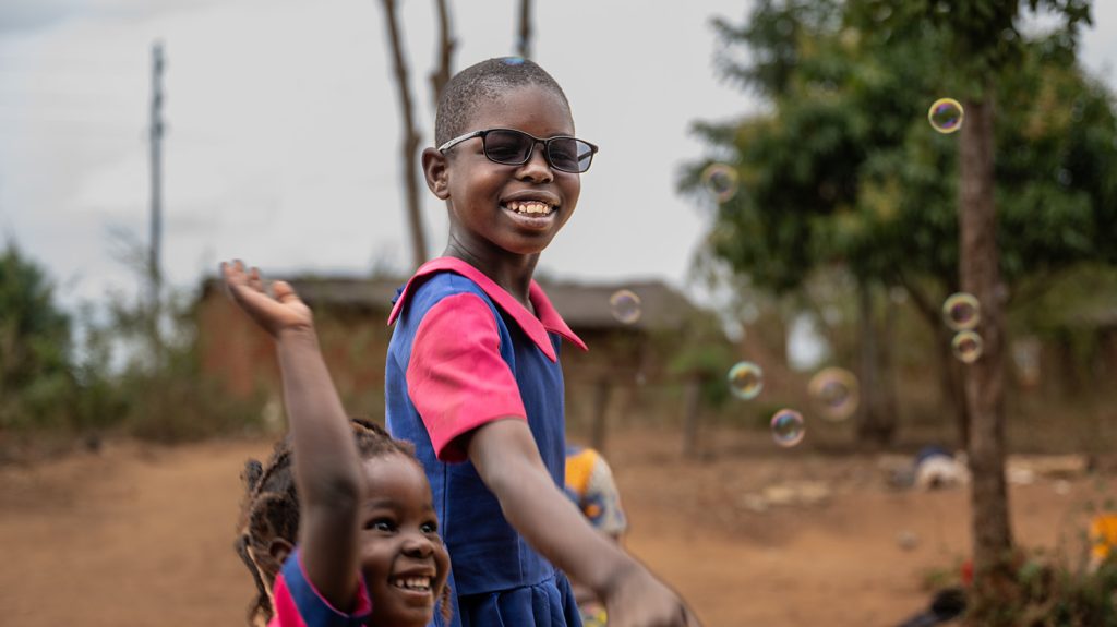 A girl smiles while wearing dark glasses. Bubbles float in the air around her. A young girl stands next to her, catching the bubbles.