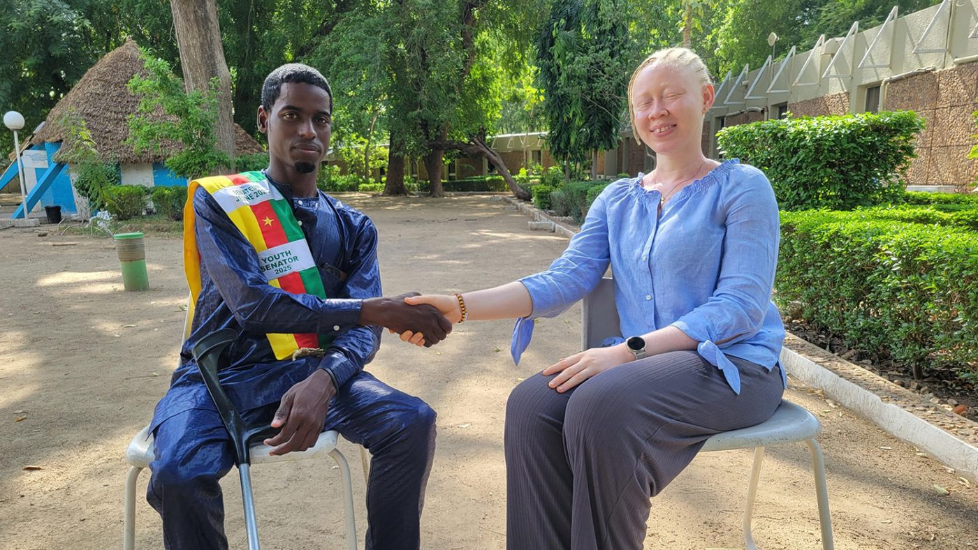 Ousmanou shakes hands with Daniella, who has albinism. Ousmanou is wearing his youth senator sash. His mobility crutch is next to him. They are both seated outside.