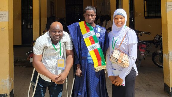 Ousmanou stands in the middle between a man using mobility crutches and a woman wearing a hijab.