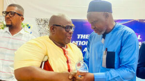 Two men shake hands during an awards ceremony. One man hands the other man an engraved glass award.