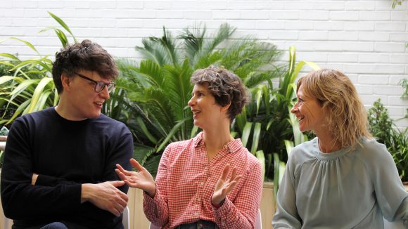 A man and two women smile and chat in an outdoor space. There are lush, green plants behind them.