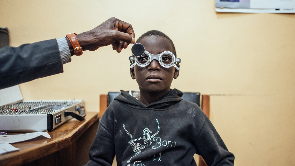 A boy sits in a chair wearing optometry glasses during an eye test. A man's hand places a lens into the right side of the pair of glasses.